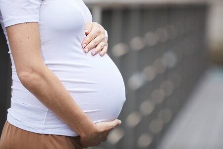Close-up of young woman in white T-shirt embracing her pregnant belly while standing on the bridge...