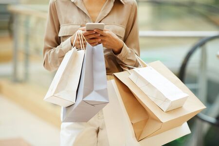 Close-up of young woman holding paper bags and communicating online on her mobile phone during...