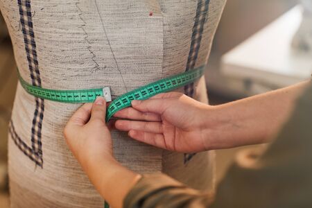 Close-up of tailor measuring the waist of mannequin with tape measure while sewing jacket in workshopの写真素材