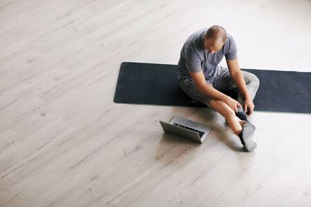 Young disabled man with prosthesis on the leg sitting on exercise mat and using laptop before trainingの写真素材