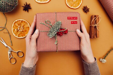 Close-up of woman holding Christmas present which she made by herself for Christmas and decorated it with fir branch and berries over yellow backgroundの写真素材
