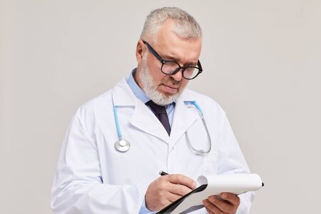 Mature doctor in eyeglasses and in white coat writing in document while standing against the white...