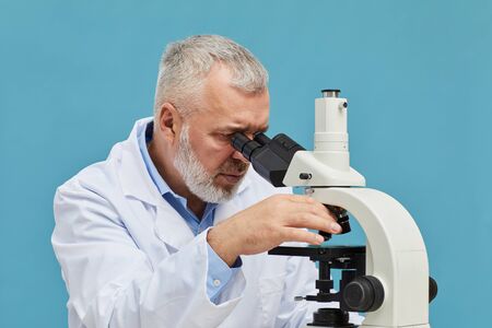 Mature scientist in white coat looking through the microscope isolated on blue background