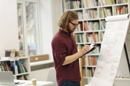Young teacher in eyeglasses standing near the whiteboard and making notes in document he preparing...