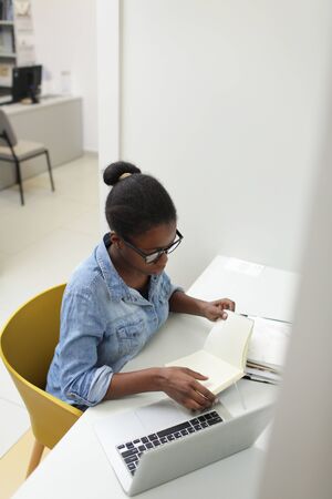 African young businesswoman in casual clothing sitting at her workplace with laptop and examining documentsの写真素材
