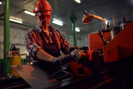 Serious mature manual worker in protective work wear standing at the lathe and making metal detailsの写真素材