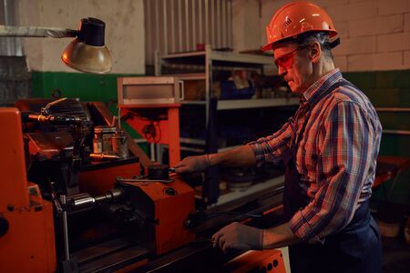 Mature engineer in protective eyesglasses and in work helmet standing near the machine and...