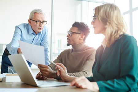 Mature businessman showing document to his colleagues and they discussing it at meeting at office