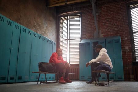 Two African men in sports clothing sitting on bench and talking to each other in locker roomの写真素材