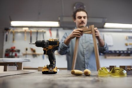 Close-up of drill and protective eyeglasses on the table with carpenter standing in the background and making wooden chairの写真素材