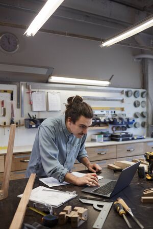 Serious young architect looking at monitor of laptop and working with blueprint at the table in...