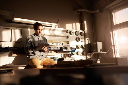 Young serious businessman sitting on his workplace with tools in the background and making notes he...