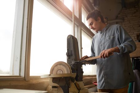 Young carpenter in casual clothing standing and cutting the wooden plank using lathe machine while...