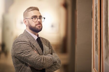 Young bearded man in eyeglasses standing with his arms crossed and looking at beautiful pictureの写真素材
