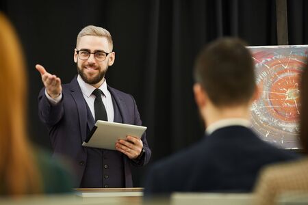 Confident businessman holding tablet pc and talking to the audience during the conference