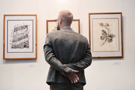 Rear view of man in suit standing in front of the wall with paintings and enjoying the art at...