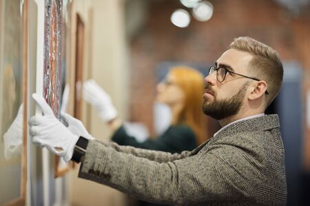 Bearded art manager in eyeglasses wearing gloves and hanging the painting on the wall with his...