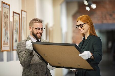 Happy man and woman carrying the big painting together they preparing for exhibition at art galleryの写真素材