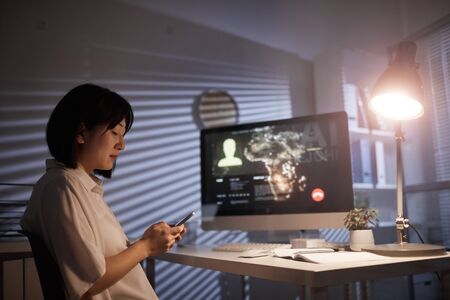 Asian businesswoman using mobile phone while sitting in front of computer monitor with online...