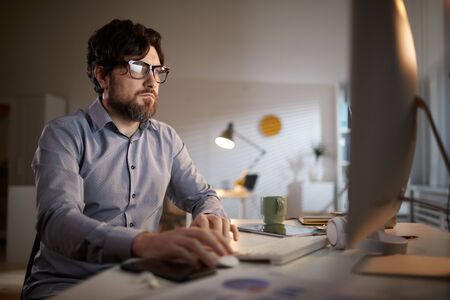 Serious bearded businessman in eyeglasses looking at computer monitor while sitting at his workplace...