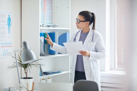 Female doctor in white coat standing near the bookcase and looking for medical cards of her patients at medical clinicの写真素材