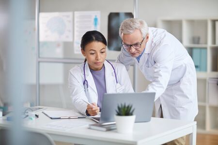 Senior doctor in white coat and young female nurse looking at monitor of laptop and discussing online presentation at officeの写真素材