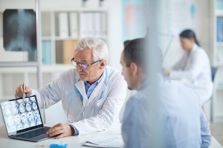 Senior doctor sitting at the table and pointing at laptop with x-ray images and explaining it to the patient during his visit at hospitalの写真素材