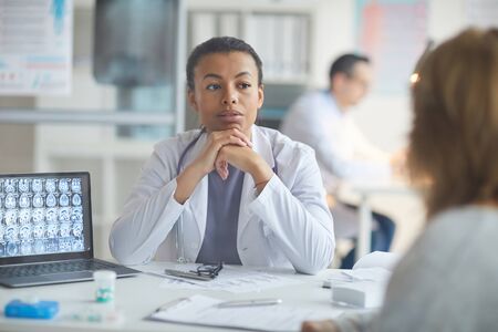 Serious female doctor sitting at her workplace and listening to complains of her patient at the hospitalの写真素材