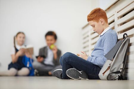 Red haired schoolboy sitting on the floor and playing in mobile phone during a break at school with other children in the backgroundの写真素材