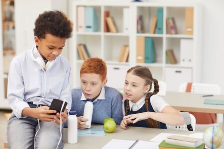 School children sitting at the table drinking milk and watching something on mobile phone during...