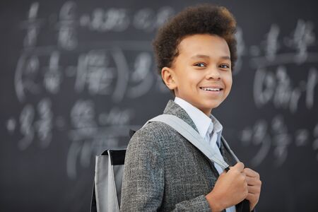 Portrait of African schoolboy standing with backpack and smiling at camera with blackboard in the backgroundの写真素材