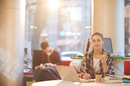 Portrait of teenage girl sitting at the table in front of laptop and looking at camera while making...