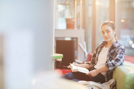 Portrait of young woman in casual clothing looking at camera while sitting on sofa and reading a...
