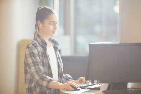 Young woman in casual clothing sitting at the table in front of computer monitor and typing on computer keyboard at officeの写真素材