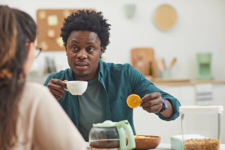 African young man drinking coffee with cookies and talking to his girlfriend in the kitchen at home