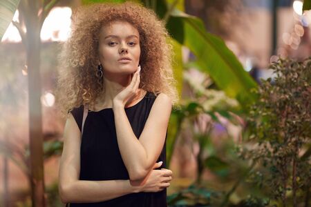 Portrait of beautiful young woman with blonde curly hair in black dress posing at camera