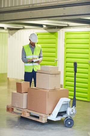Young man in working clothing making notes in document while standing near the cart with boxes in...