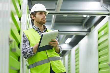 Serious young manual worker in working clothing standing and working with documents in warehouseの写真素材
