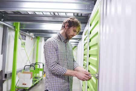 Young bearded man in casual clothing standing and opening storage box in warehouseの写真素材