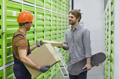 Young worker in work helmet giving a parcel to young man while they standing in stockroomの写真素材