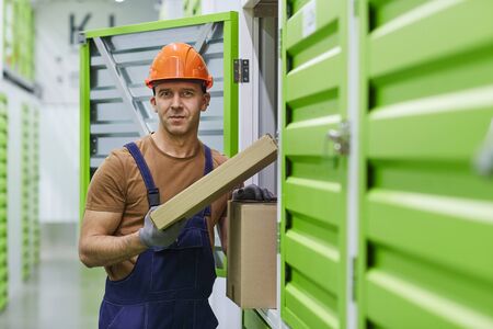 Portrait of young worker in work helmet and in overalls looking at camera while putting parcels into the boxes in warehouseの写真素材
