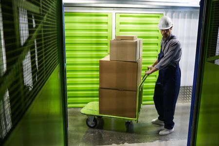 Young man in work helmet and in overalls delivering the parcels he carrying cart with stack of boxes