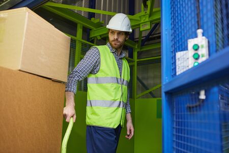 Young worker in protective workwear carrying boxes on cart while working in stockroom