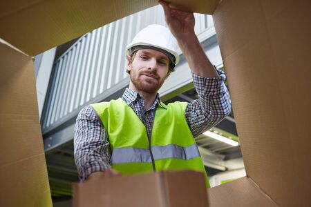 Young manual worker in work helmet packing or unpacking cardboard box in warehouse