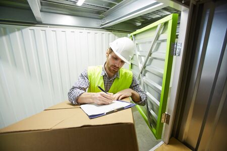 Young foreman in work helmet writing in document and making a parcel before shipping in warehouseの写真素材
