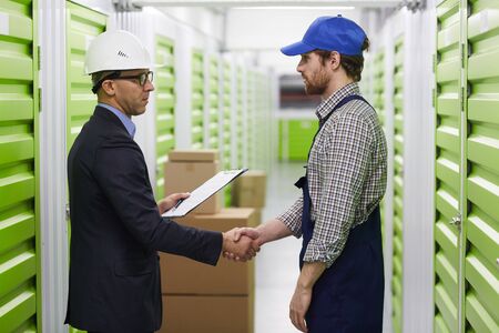 Serious businessman in work helmet shaking hand to worker in overalls while they standing in storage...