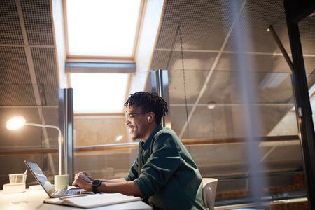 African young designer sitting at his workplace typing on laptop computer and smiling during work...