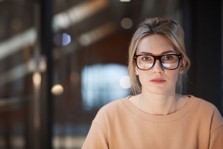 Portrait of young woman with blonde hair and in eyeglasses looking at camera while sitting at officeの写真素材