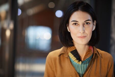 Portrait of young businesswoman with dark hair and in wireless headphones looking at camera while working at officeの写真素材