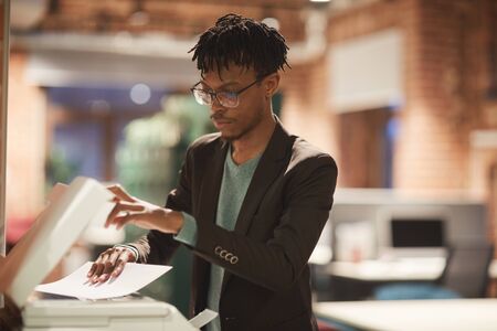 Young African businessman in eyeglasses standing near the printer and making copy of documents at...